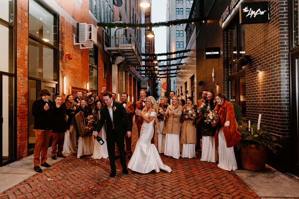  bride in a trumpet style gown and the groom in a green suit, the groomsmen in caramel notch lapel suits and the bridesmaids in ivory gowns 