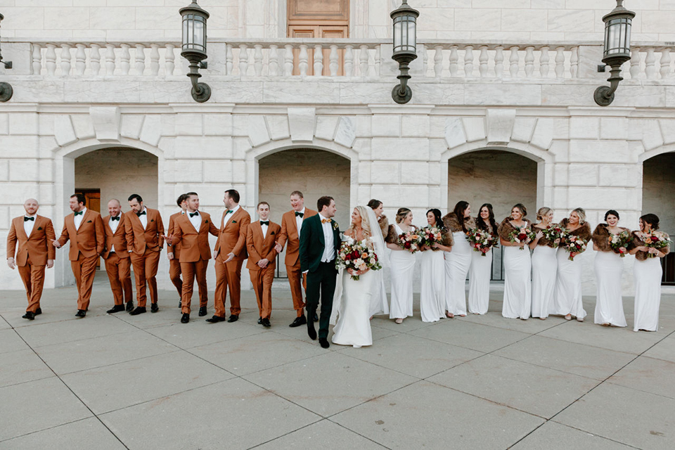  bride in a trumpet style gown and the groom in a green suit, the groomsmen in caramel notch lapel suits and the bridesmaids in ivory gowns 