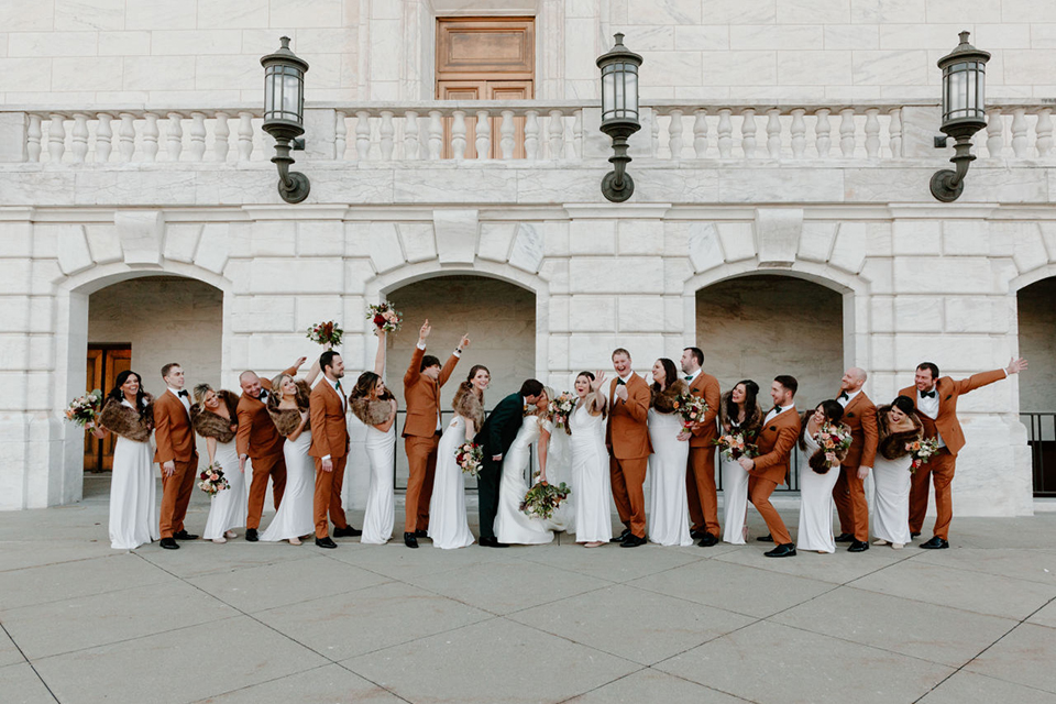  bride in a trumpet style gown and the groom in a green suit, the groomsmen in caramel notch lapel suits and the bridesmaids in ivory gowns 