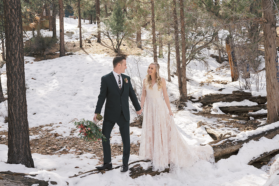  big bear elopement in the snow with the bride is a lace gown and the groom in a green suit – couple walking on snowy path 