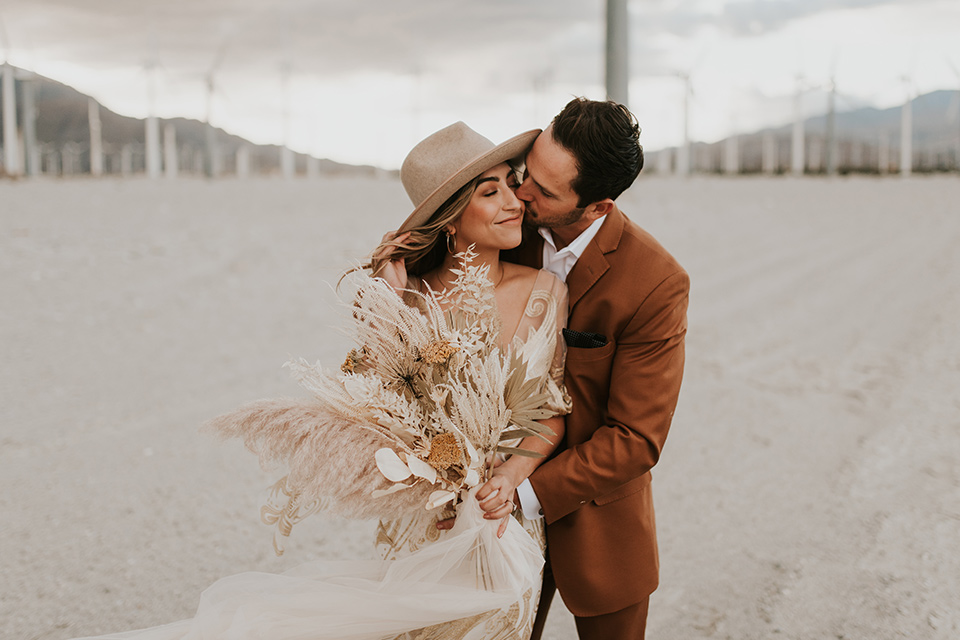  eloping in the desert with a bohemian vibe – bride in a lace gown and the groom in a caramel brown suit – groom kissing bride on the cheek 