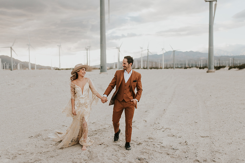  eloping in the desert with a bohemian vibe – bride in a lace gown and the groom in a caramel brown suit – couple walking