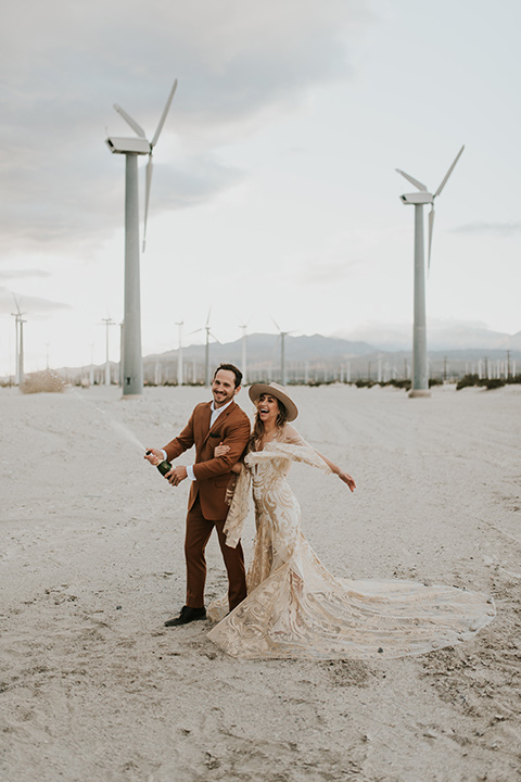  eloping in the desert with a bohemian vibe – bride in a lace gown and the groom in a caramel brown suit -couple popping champagne