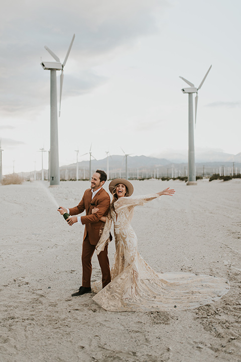  eloping in the desert with a bohemian vibe – bride in a lace gown and the groom in a caramel brown suit - couple popping champagne 