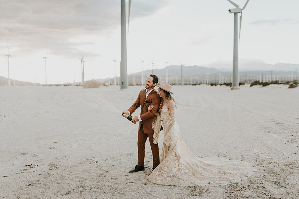  eloping in the desert with a bohemian vibe – bride in a lace gown and the groom in a caramel brown suit – popping champagne