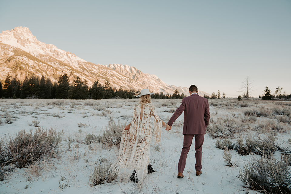  grand tetons elopement in the winter with snow on the ground with the bride in a lace gown and the groom in a rose pink suit – couple walking in the snow 
