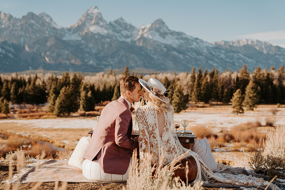  grand tetons elopement in the winter with snow on the ground with the bride in a lace gown and the groom in a rose pink suit – couple sitting at a picnic 