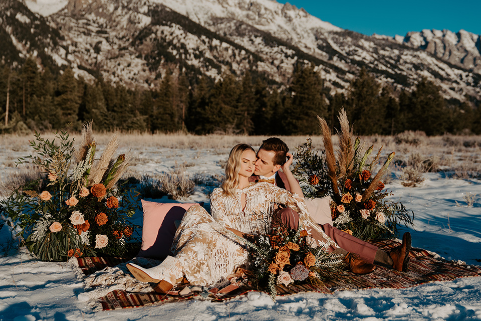  grand tetons elopement in the winter with snow on the ground with the bride in a lace gown and the groom in a rose pink suit – couple sitting at a picnic 