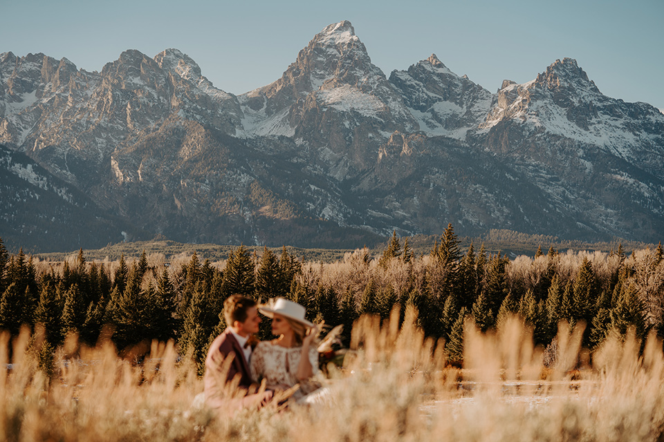  grand tetons elopement in the winter with snow on the ground with the bride in a lace gown and the groom in a rose pink suit – couple sitting at a picnic 