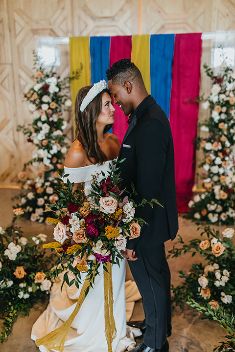  modern and colorful wedding with the bride in an off-the-shoulder dress and the groom in an all-black tuxedo – couple exchanging vows 