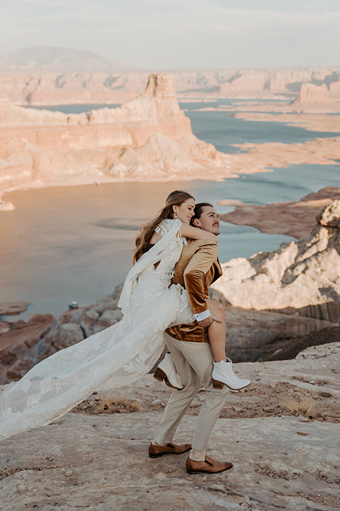  a boho wedding at lake powell with the bride in a bohemian gown and a wide brimmed hat and the groom in a gold velvet jacket – bride on groom’s back