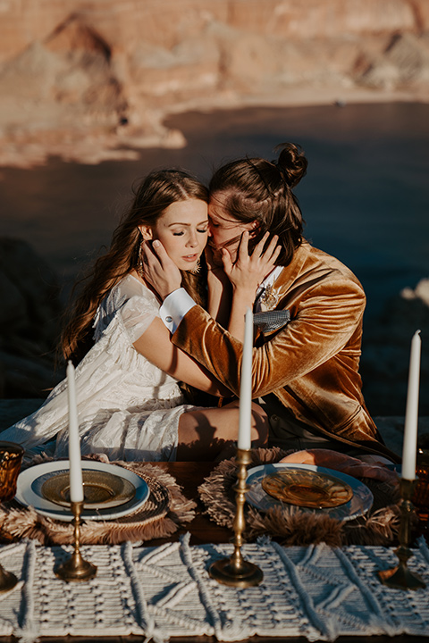  a boho wedding at lake powell with the bride in a bohemian gown and a wide brimmed hat and the groom in a gold velvet jacket – couple sitting and kissing