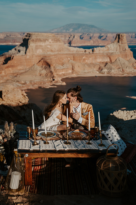  a boho wedding at lake powell with the bride in a bohemian gown and a wide brimmed hat and the groom in a gold velvet jacket – couple sitting and kissing 