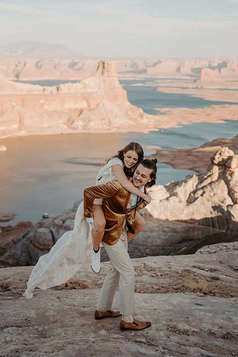  a boho wedding at lake powell with the bride in a bohemian gown and a wide brimmed hat and the groom in a gold velvet jacket – bride on groom’s back 